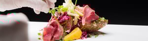 Ein kunstvoll angerichtetes Gourmetgericht mit dünn geschnittenem Fleisch, Brot, Gemüse und Obststücken, das von einer behandschuhten Hand mit Kräutern garniert wird.