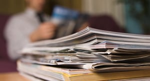 A stack of magazines lies on a table in the foreground, while a person is seen reading a magazine in the blurred background.