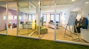 Blick durch eine Glaswand in ein modernes Büro mit mehreren Arbeitsplätzen, einem Flipchart, einem Anzug auf einem Kleiderständer und grünem Teppichboden im Vordergrund.