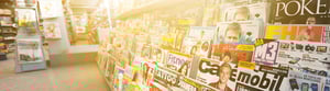 Magazine rack in a kiosk or store, filled with colorful magazines on topics such as celebrities, lifestyle, technology, and fitness, lit by warm sunlight.