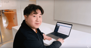 Person sitting at a white table in a modern office, working on a laptop. A web application is open on the screen. The person is looking at the camera with a friendly expression. In the background, there's a room with a glass wall and an orange standing table.