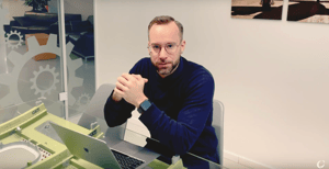 Mann mit Brille und Smartwatch sitzt an einem Tisch mit Laptop und Modellteilen, in einem modernen Büro mit Zahnradsymbolen an der Glaswand.
